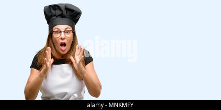 Middle age cook woman wearing chef apron stressful keeping hands on head, terrified in panic, shouting isolated blue background Stock Photo
