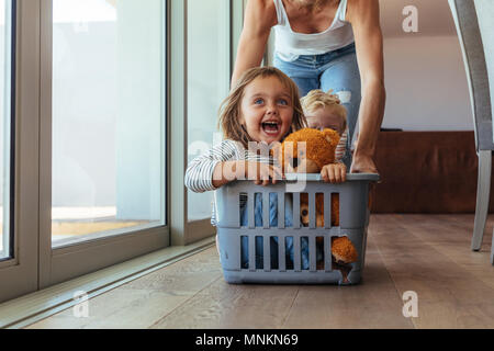 Excited little girl with teddy bear and her brother sitting in a laundry basket being pushed by her mother. Cute family playing together at home. Stock Photo
