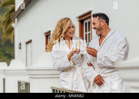 Beautiful couple in bathrobe clinking glasses of wine and smiling. Cheerful couple wearing bathrobes with glasses of champagne in luxury villa. Stock Photo