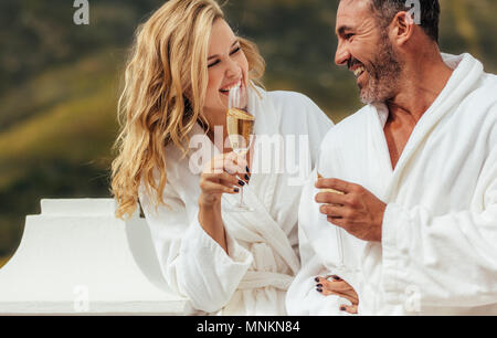 Cheerful couple in bathrobe talking and laughing in the balcony with glass of wine. Man and woman in bathrobes spending time at together. Stock Photo