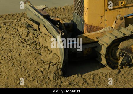 Close-up, detail, blur, scraper blade of old yellow construction bulldozer pushing crumbling wet sand, motion Stock Photo