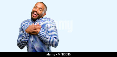 African american man with beard having charming smile holding hands on heart wanting to show love and sympathy isolated over blue background Stock Photo