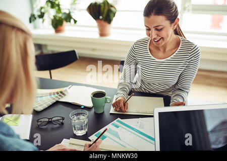 Two smiling young businesswomen sitting at a table in an office going over documents together Stock Photo