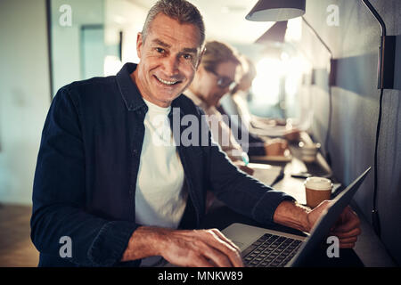 Smiling mature designer working on a laptop while sitting in a row with coworkers at a table in a modern office Stock Photo
