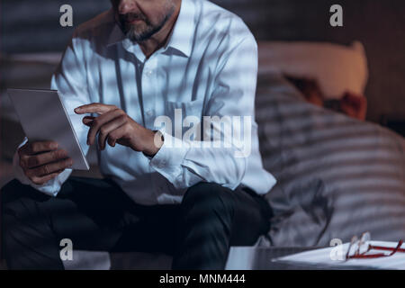 Low angle view of a mature male sitting on a bed in a room and holding a tablet in his hand with a woman sleeping under bed-cover in a blurred backgro Stock Photo