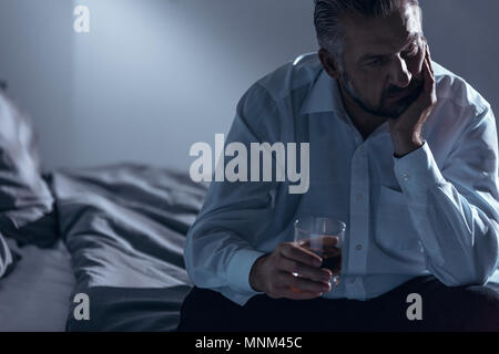 Close-up of a man with midlife crisis sitting on a bed with one hand on his chin and holding a glass of alcohol in the other Stock Photo