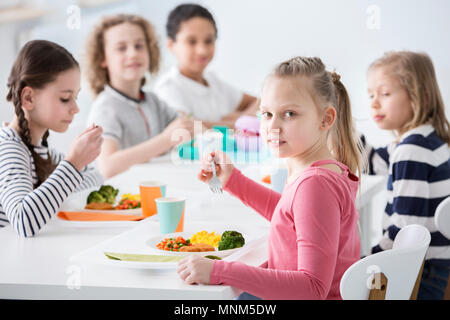Girl eating vegetables with friends in the canteen during break at school Stock Photo