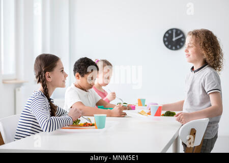 Happy boy and friends sitting at table with healthy food at school, in dining hall Stock Photo