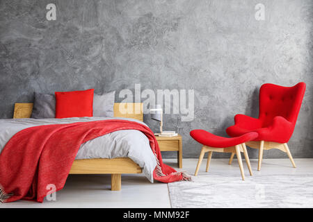 Red armchair and stool next to wooden bed against concrete wall with copy space in bedroom Stock Photo