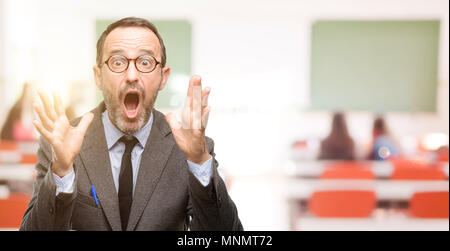 Teacher man using glasses stressful keeping hands on head, terrified in panic, shouting at classroom Stock Photo