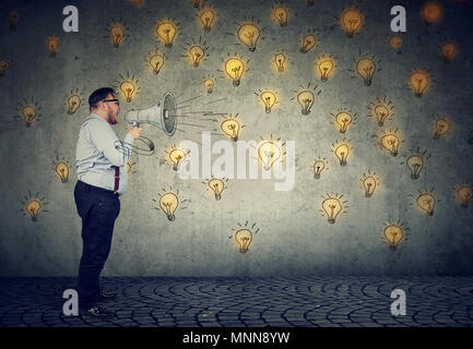 Man screaming out his ideas loud in megaphone spreading his ideas isolated on gray wall background Stock Photo