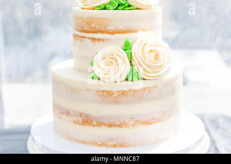 Macro closeup of three tiered vanilla golden light white yellow cream sponge wedding cake with rose flowers decoration on bright stand, green leaves Stock Photo