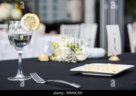 Closeup of one glass with lemon slice and water in restaurant or wedding reception black table tablecloth, plate with cake slice, bouquet flowers, for Stock Photo