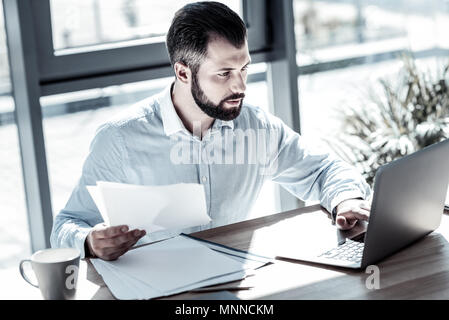 Concentrated serious man holding the document and using the laptop. Stock Photo