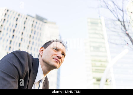 Handsome, attractive young businessman sitting smiling looking up in suit and tie, cheerful on interview break in urban park, skyscrapers Stock Photo