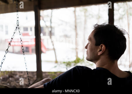 Closeup of young man sitting on swing under wooden deck of house on backyard in neighborhood with snow during blizzard white storm, snowflakes falling Stock Photo