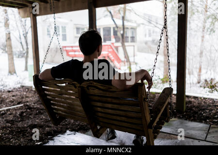 Young man sitting on swing under wooden deck of house on backyard in neighborhood with snow during blizzard white storm, snowflakes falling in Virgini Stock Photo