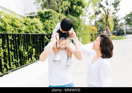 Cute Asian father piggybacking his son along with his wife in the park. Excited family spending time together with happiness Stock Photo