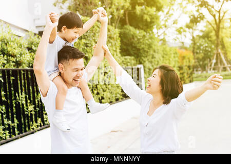 Cute Asian father piggy backing his son along with his wife in the park. Excited family raising hands together with happiness Stock Photo