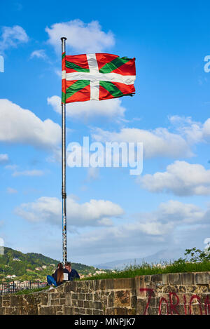 Two young people sitting under the mast of a Ikurrina, Basque Country flag waving on a blue sky in the Monte Urgull of San Sebastian. Spain. Stock Photo
