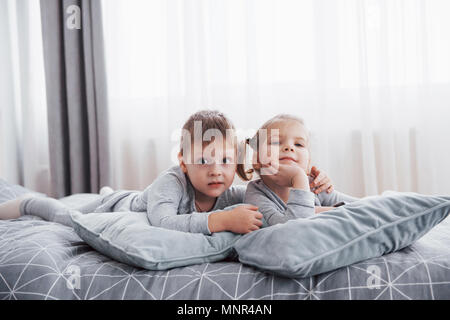 Happy kids playing in white bedroom. Little boy and girl, brother and sister play on the bed wearing pajamas. Nursery interior for children. Nightwear and bedding for baby and toddler. Family at home Stock Photo
