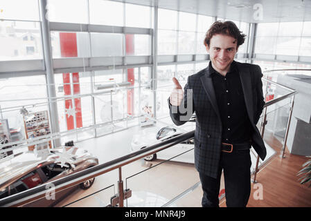 Visiting car dealership. Young man on car salon background. Happy man shows finger up. Car sale and rent concept Stock Photo