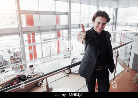 Visiting car dealership. Young man on car salon background. Happy man shows finger up. Car sale and rent concept Stock Photo