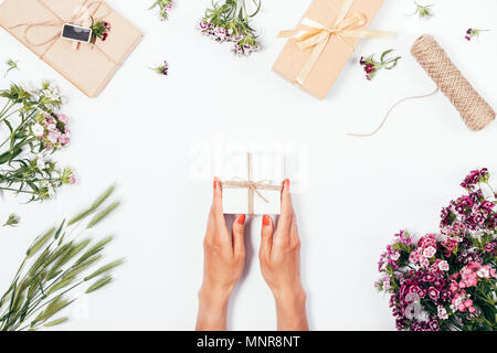 Top view of female hands with red nail polish hold a small gift box among summer flowers and presents. Festive flat lay frame on a white background. Stock Photo