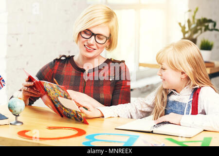 Successful kid girl studying English language with elegant blonde female teacher, table with books and letters, learning process in white stylish classroom Stock Photo
