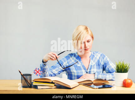 Smart female student, blonde woman holds eyeglasses in hand, studying English language on table with books, successful learning concept Stock Photo