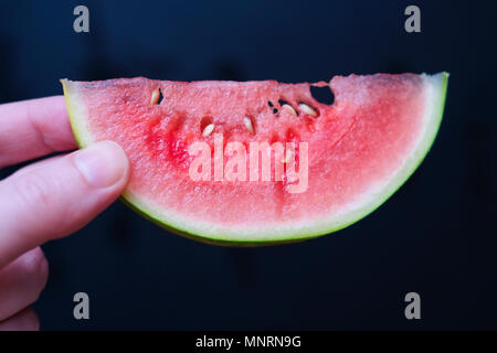 A slice of juicy fresh mini watermelon in a female hand on a dark background. Selective focus. Stock Photo