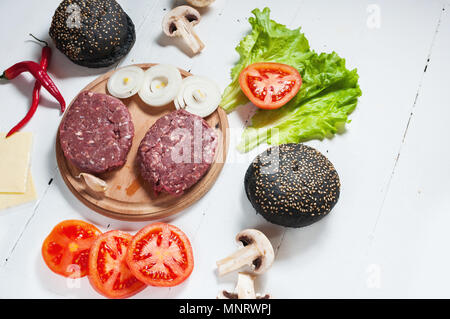 Homemade hamburger ingredients. Raw minced beef, fresh black bun, slice of cheese, tomato, onion rings, lettuce on wood white background. Stock Photo