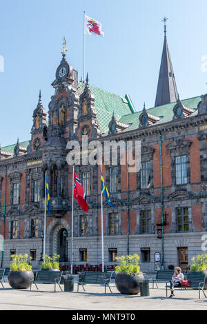 Woman sites on metal bench outside Malmo town hall facade with flag flying above clock tower and Swedish flag on flagpole at front of building. Stock Photo