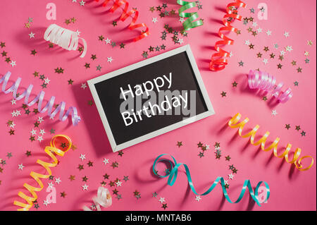 Colorful party streamers, gold little stars and blackboard with text Happy Birthday on pink backgrond. Stock Photo