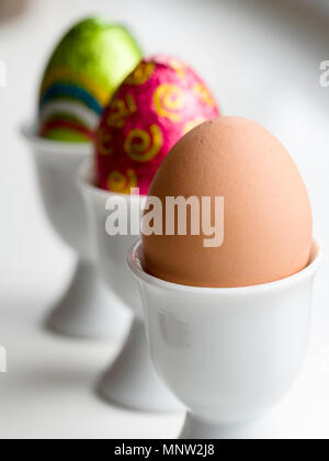 Chocolate Easter Eggs and a brown hen's egg in egg cups: A row of three eggs in three white porcelain egg cups on a white background. Stock Photo
