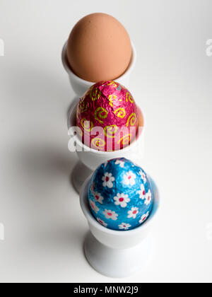 Chocolate Easter Eggs and a brown hen's egg in egg cups: A row of three eggs in three white porcelain egg cups on a white background. Stock Photo