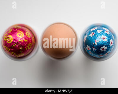 Chocolate Easter Eggs and a brown hen's egg in egg cups: A row of three eggs in three white porcelain egg cups on a white background. Stock Photo
