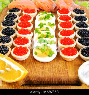 Homemade tartlets with red and black caviar, salmon, lettuce and eggs. On a cutting board. Concept of luxury food. Square. Close-up. Stock Photo