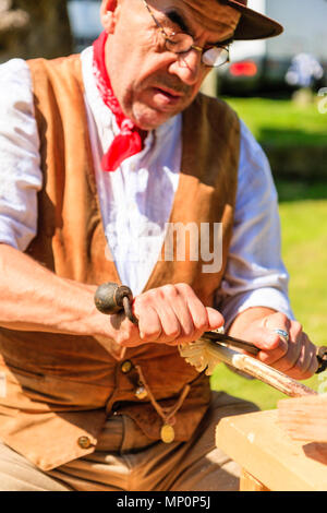 Salute to the 40s popular event. Carpenter, woodworker in 1940's country clothing using a drawknife to shave a wooden pole held on workbench. Stock Photo