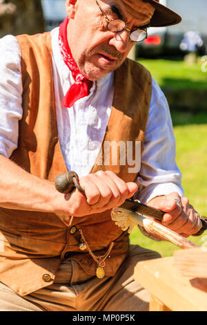 Salute to the 40s popular event. Carpenter, woodworker in 1940's country clothing using a drawknife to shave a wooden pole held on workbench. Stock Photo