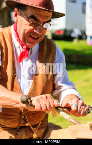 Salute to the 40s popular event. Carpenter, woodworker in 1940's country clothing using a drawknife to shave a wooden pole held on workbench. Stock Photo
