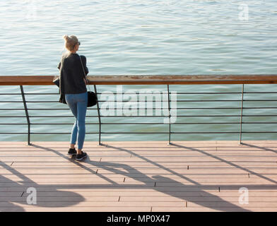 Belgrade, Serbia - October 15, 2017 : Young blonde woman standing  on the river promenade leaning on the railings and looking at water, from behind Stock Photo