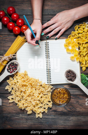 Female hands writing on a notepad. spices, pasta and vegetables around notebook on a wooden table. Mockup. Stock Photo