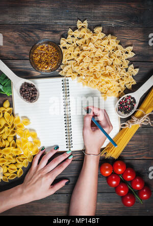 Female hands writing notepad. spices, pasta and vegetables around notebook on wooden table. Mockup. Stock Photo