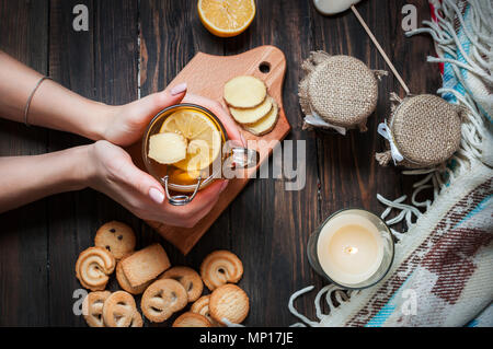 female hands holding cup black tea with lemon, ginger and cookies on dark wood. Stock Photo