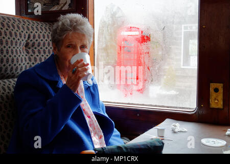An older woman drinking tea on the Peak Rail steam train on a rainy day in in Bakewell, Derbyshire with a red phone box outside Stock Photo