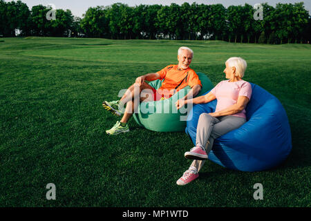 Senior people, elderly man and woman sitting on beanbags on grass, looking at each other and laughing, Stock Photo