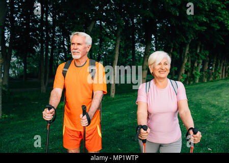 elderly couple with trekking poles hiking below trees in woods. Stock Photo