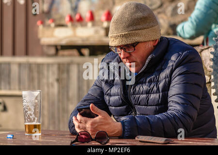 Man in warm coat and hat sat outside a pub with a nearly finished pint of beer and using a mobile phone. Teignmouth, Devon. Feb 2018. Stock Photo