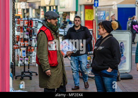 Woman talking in a friendly manner to a man selling Big Issue magazines on a high street, Teignmouth, Devon. Feb 2018. Stock Photo
